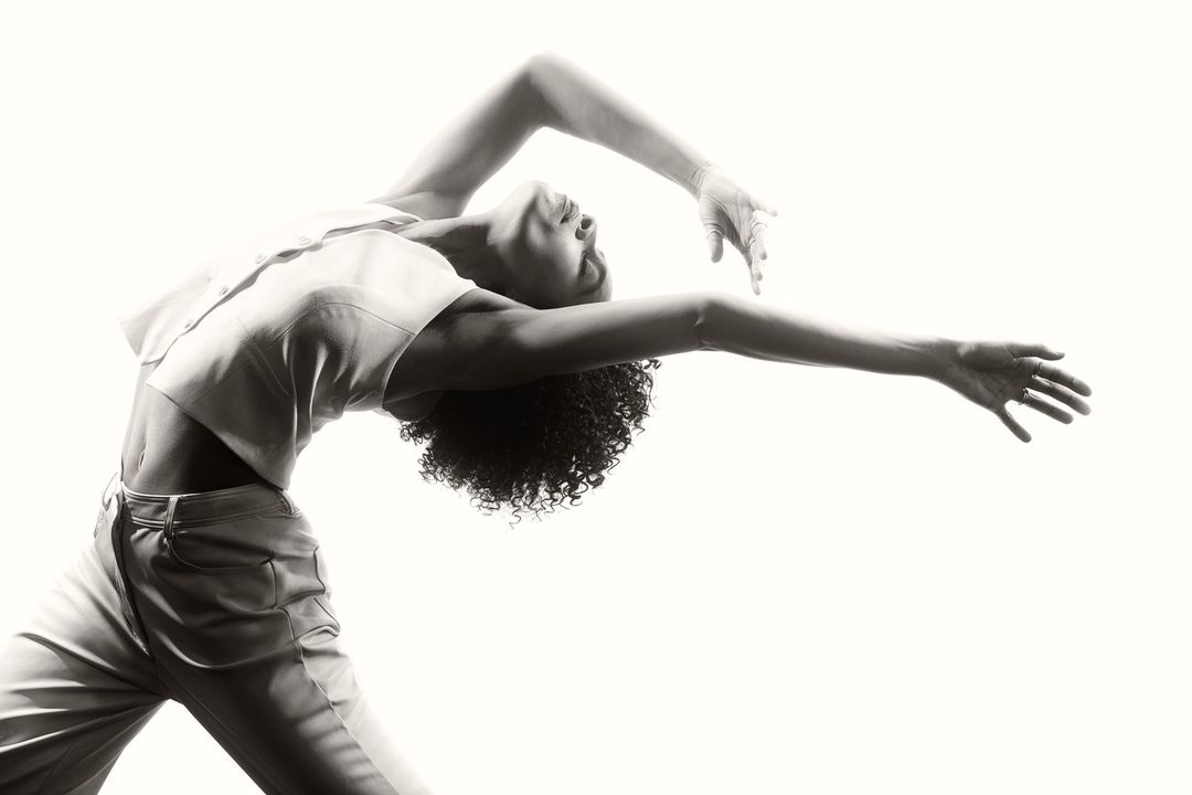 Artistic black and white studio portrait of a woman in motion wearing a crop top and pants against a bright white background.